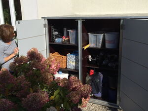 . Ein offener Gartenschrank mit Gartenzubehör und Behältern, daneben eine Frau und blühende Hortensien.