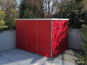 . Ein rotes Gartenhaus steht auf einer gepflasterten Terrasse, umgeben von Bäumen und Sträuchern.