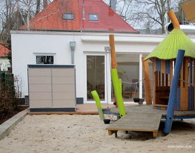 . The image shows a playground area with a sandbox in the foreground. There is a play structure that resembles a small house with colorful poles and a green roof. It's situated on a wooden platform with a small ramp leading up to it. In the background, there's a modern-looking building with large windows and a red roof. There's also a shed-like structure next to the building. Trees are visible around the area.