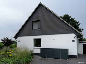 . The image shows the exterior of a house with a triangular roof. The house has white brick walls and dark shingles on the roof. There are some plants with yellow flowers in the garden area in front of the house. There are also three dark-colored storage units or containers next to the house on a paved area. A drainpipe is visible on the right side of the house, and there's a window with a curtain on the ground floor.
