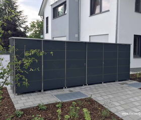 The image shows a large, dark-colored outdoor storage unit or shed next to a modern house. The structure is positioned on a paved area, surrounded by landscaped garden beds with mulch and plants. The background includes a white and gray house with windows.