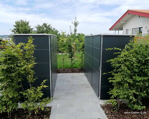 . The image shows two dark-colored garden storage units outside, surrounded by green shrubs. They are positioned on either side of a paved pathway, leading to a lawn area in the background. There's a house with a red roof visible on the right side. It looks like a neatly maintained garden area.