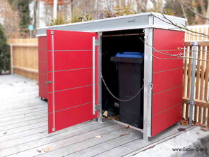 . The image shows a red outdoor storage unit with open doors. Inside, there's a large garbage bin with a blue lid. The unit has a flat roof with plants on top, and it's placed on a deck or patio. There's a chain visible near the open doors, and a wooden fence is in the background.