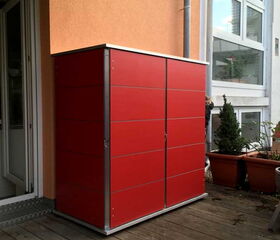 . The image shows a bright red storage unit or cabinet situated on a wooden deck. It is placed against the wall of a building, next to a door. There are some potted plants nearby. The storage unit appears to be outdoors, likely used for organizing garden tools or other items.