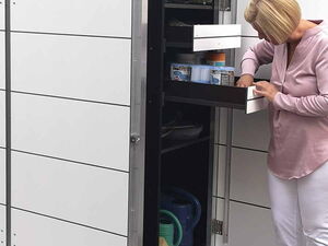 . The image shows a woman interacting with a tall storage unit or cabinet, which is located outdoors. The cabinet has several drawers and compartments, some of which seem to hold gardening tools and supplies like watering cans and containers. The top of the cabinet appears to have plants on it. The woman is inspecting the contents of one of the open drawers.