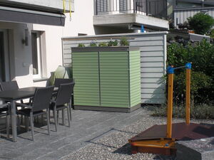 . The image shows a patio area with several objects:

1. A dining table with several chairs.
2. A large green storage unit or cabinet with plants on top.
3. A structure with two wooden posts and a horizontal bar, possibly an outdoor gym or play equipment.
4. Surrounding greenery, fencing, and a balcony above.
5. A stone or tiled floor with a section of gravel.