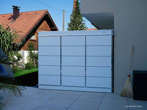 . The image shows a white metal storage unit or cabinet located outdoors. It's placed against a wall on a patio or tiled surface. There's a broom leaning against the wall nearby, and part of a chair or similar object is visible. In the background, there is a wooden house with a red roof and some greenery.