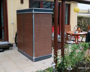 . Holzschuppen auf Terrasse neben Tisch mit Stühlen und Pflanzen im Gartenbereich.