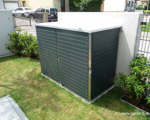 The image shows a garden storage unit, possibly made of metal or synthetic material, with a sleek, modern design. It is placed on a grassy area next to a concrete wall and surrounded by some small plants. The unit appears to be locked or secured, and the environment looks like a residential area with a driveway and cars visible in the background.