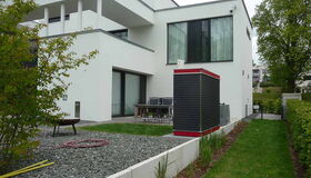 . This image shows a modern white house with a flat roof. There is a garden area with gravel and grass, along with some shrubs or small trees. A patio area with chairs and a table is also visible. In the foreground, there's a tall, narrow structure with a red top, possibly a storage unit or utility box.