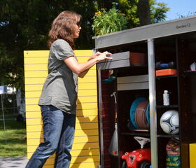 . The image shows a person interacting with a storage shed or cabinet labeled "Garten Q." The cabinet is outdoor, likely used for garden or yard equipment. Visible items include a ball, a toy vehicle, a broom, a hose reel, and various garden tools and supplies. There are plants on top of the cabinet, and the person appears to be accessing or organizing items within it.