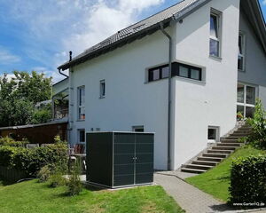 . Ein modernes Haus mit Garten, Treppe, Rasen und einem großen schwarzen Schrank im Vordergrund.