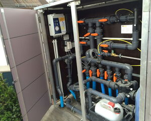 The image shows a technical enclosure or cabinet containing a complex system of pipes and valves, likely part of a pool or water treatment setup. It features:

1. Multiple gray pipes with orange-handled valves.
2. A white control box with dials and indicators.
3. Various connectors and fittings, including blue tubing.
4. Electrical wiring and cable management.
5. A white plastic container, possibly for chemicals or maintenance solutions. 

The system appears to be organized for efficient flow control and maintenance access.