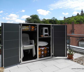 . The image shows a storage cabinet on a rooftop terrace. It contains several stacked white plastic chairs, folded towels or blankets, and some other items like a folded stool and a basket. The cabinet doors are open, and there are plants in pots placed around the terrace. The background shows rooftops and a building on a hill.