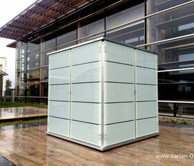 . The image depicts a modern, cube-shaped structure with frosted glass panels and metallic frames. It's placed outdoors on a tiled surface, next to a contemporary building with large glass windows. The structure appears to be a garden storage unit or similar architectural element.