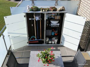 . Ein Balkon mit offenem Schrank voller Gartengeräte, Pflanzen oben und Tisch mit Blume davor.