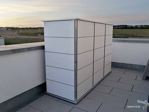 . Ein weißer Gartenschrank auf einer Terrasse, im Hintergrund sind Felder zu sehen.