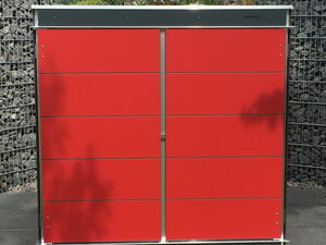 . The image shows a modern red storage unit, likely made for outdoor use. It is placed on a tiled surface with grass in the foreground. Behind it, there's a wall made of wire cages filled with rocks. The roof of the storage unit has greenery or plants growing on it. The setting appears to be an outdoor garden or patio area.