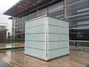 . The image shows a cubical structure with frosted glass panels and metal edges, set on what appears to be a wooden platform outdoors. It is located near a modern building with large glass windows and metal accents. The setting looks contemporary and architectural, possibly an outdoor or urban area.