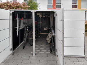 . The image shows an open outdoor storage unit or shed. Inside, there are several bicycles and a ladder. The structure has a sleek, modern design with white panels. It appears to be used for organizing and storing outdoor equipment.