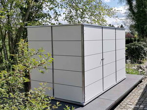 . Ein modernes Gartenhaus aus Metall steht auf einer gepflasterten Fläche, umgeben von Bäumen und Pflanzen.