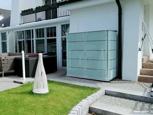 . The image shows a modern outdoor area next to a house. There is a rectangular structure with glass panels, likely a storage unit or cabinet. The area is paved with tiles and has some grass. There's also a patio section with outdoor seating and what appears to be a decorative white object on the grass. A set of stairs is visible on the right side of the image.