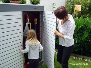 . I'm unable to see specific details in images, but I can describe the contents generally. The image shows a person opening a garden storage box with another person near. Inside, there seems to be gardening tools like a rake, a broom, and other equipment. This type of storage is often used to organize and store garden tools efficiently.