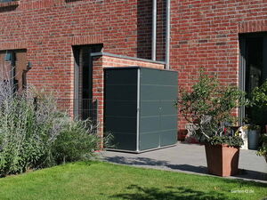 . Ein graues Gartenhaus steht auf einer Terrasse neben einer Ziegelwand, umgeben von Pflanzen und Rasen.