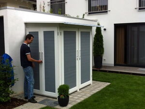 . Ein Mann öffnet eine Gartenhütte neben einem modernen Haus mit Terrasse und Garten.