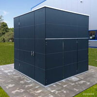 The image shows a modern, dark-colored storage shed or outdoor structure, placed on a paved area with grass and trees in the background. The shed has a minimalist, sleek design with clean lines and a flat roof. There is a handle on one of the panels, indicating a door.