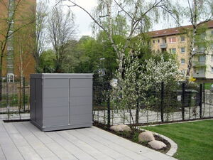 . The image shows a grey metal storage shed located in a landscaped area. It is surrounded by paving stones and situated next to a fence. There are trees, shrubs, and flowering plants around the shed, and an apartment building is visible in the background. The setting appears to be an outdoor residential area with well-maintained greenery.