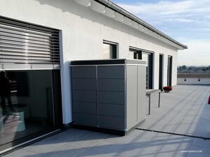 . Ein grauer Schrank steht auf einer modernen Terrasse neben einem weißen Gebäude mit großen Fenstern.