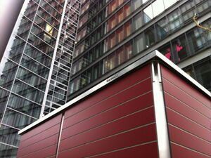. The image shows a modern architectural scene with a focus on a red panel structure in the foreground. Behind it, there is a tall, glass-fronted building with multiple windows. The urban setting suggests a city environment, and the structure has a metal frame. The words "Garten Q" are visible on the structure.