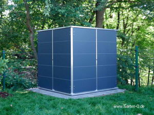 . The image depicts a modern, cube-shaped structure or storage unit placed outdoors on a concrete slab. It is surrounded by grassy lawn and trees, suggesting a garden setting. The unit has a sleek, dark blue surface with metallic edges. The website "www.Garten-Q.de" is visible in the bottom right corner.