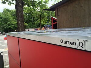. Ein rotes Gartenhaus von Garten[Q] im Freien, mit Bäumen und einem Spielplatz im Hintergrund.