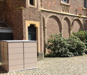 . The image depicts a tall, rectangular storage unit placed in front of a historical brick building with arched windows. The building has ornate detailing, and there is greenery, such as bushes, alongside the stone-paved ground.