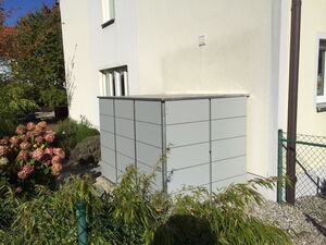 . Ein moderner, grauer Außenaufbewahrungsschrank steht an der Seite eines Hauses, umgeben von Gartenpflanzen.