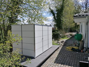 Ein weißer Schuppen neben einem Haus, umgeben von Bäumen und Gartenwerkzeug auf gepflastertem Weg.