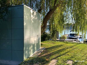 . Ein moderner Gartenschuppen steht unter einem Baum neben einem See mit einem Bootssteg.