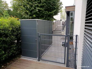 . Ein grauer Metallzaun mit Tor, dahinter ein Gartenschrank, daneben eine Treppe und grüner Bewuchs.