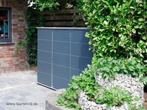 . Ein grauer Gartenschrank neben einer Mauer und einem Zaun mit grünen Pflanzen und Pflastersteinen.