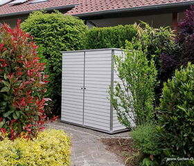 . The image features a garden scene with a small, modern shed or storage unit, possibly made of metal or wood with a metallic finish, placed among various types of lush greenery and shrubs. There are numerous plants surrounding the shed, creating a dense, vibrant setting. A house with a tiled roof is visible in the background.