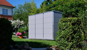 . The image depicts an outdoor garden scene featuring a large, gray storage shed. The shed is situated within a landscaped area, surrounded by well-maintained bushes and flowers. There is a house visible in the background, along with various trees and plants. The setting appears to be a residential garden space.