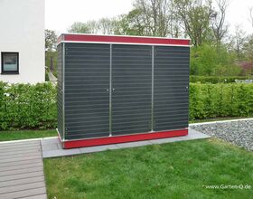 The image shows a garden storage unit with a dark exterior and a red base. It's positioned on a paved area next to a patch of grass. There are hedges and a building with a small window in the background, and trees are visible in the distance.