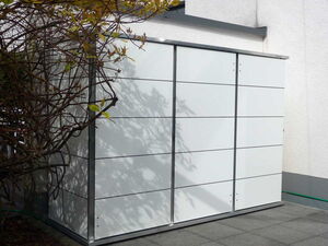 . The image shows an outdoor storage shed with a sleek, modern design. It has a flat roof and smooth, panel-like surfaces. The shed is positioned against a wall, and there are tree branches with leaves casting shadows on its surface. The setting appears to be a patio or a paved area.