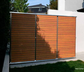 . This image shows a storage unit or shed with a modern design featuring horizontal wooden slats and metallic framing. It is set outdoors, with some grass in the foreground. There's also a house and some trees visible in the background. The unit appears to be a part of a garden setup, as indicated by the branding.