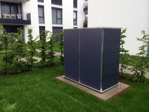 . Ein rechteckiger, grauer, schrankähnlicher Kasten steht im Gras zwischen Bäumen vor einem Wohngebäude.