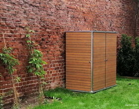 Ein Gartenschrank aus Holz steht auf Rasen vor einer Ziegelmauer, neben einigen Pflanzen.