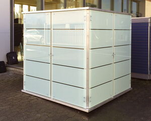 Eine überdachte Box mit durchscheinenden Seiten und Metallrahmen steht draußen neben einem Gebäude.
