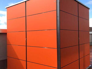. The image shows a modern-looking storage unit or shed with a sleek design. It's made of sections, with panels painted in bright orange, and it features metallic edges. The structure appears to be placed on a wooden deck with a clear sky in the background.